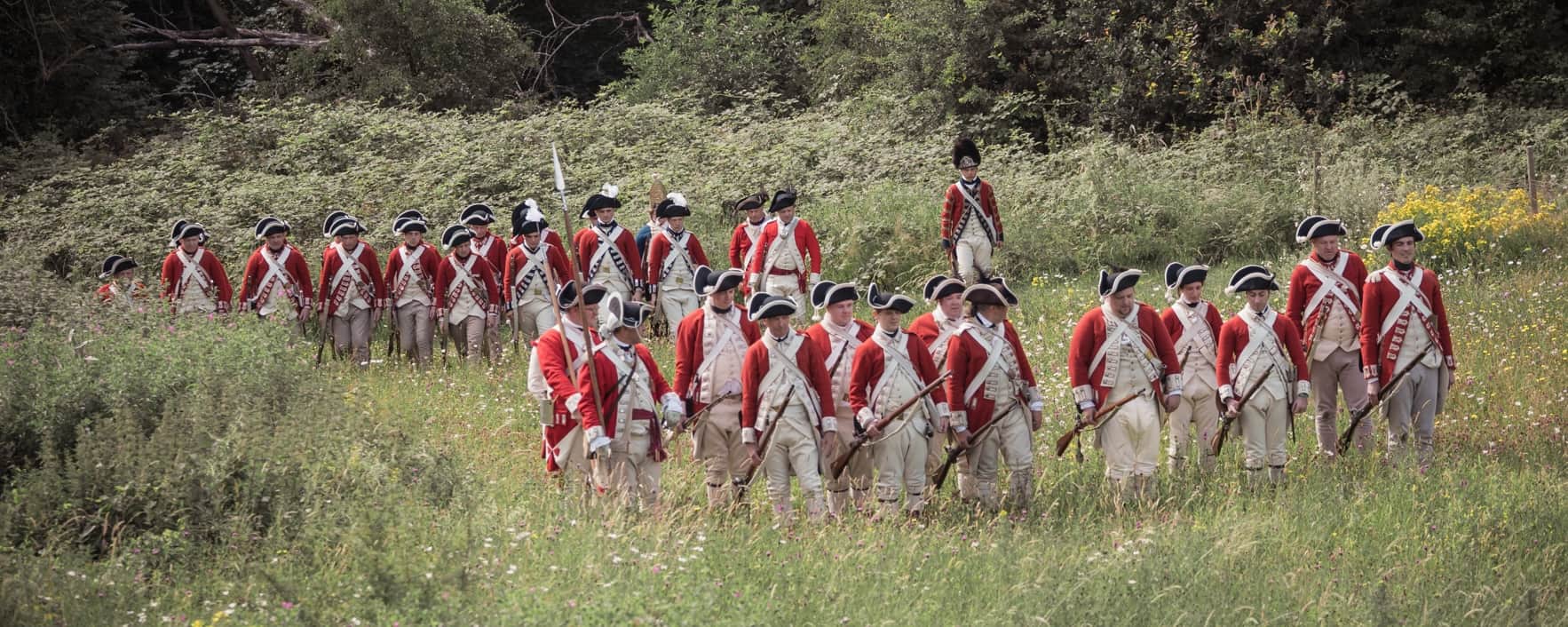 A company of late 18th century British soldiers in red coats advance across a meadow.